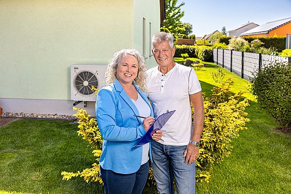 Energieberaterin Frau Jusczyk im Gespräch mit einem Kunden Eine Energieberaterin der Stadtwerke Merseburg GmbH und ein Mann stehen vor einem Haus mit Wärmepumpe und lächeln in die Kamera.