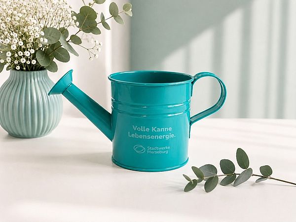 Türkisfarbene Mini-Gießkanne mit Aufschrift "Volle Kanne Lebensenergie" neben einer Blumenvase