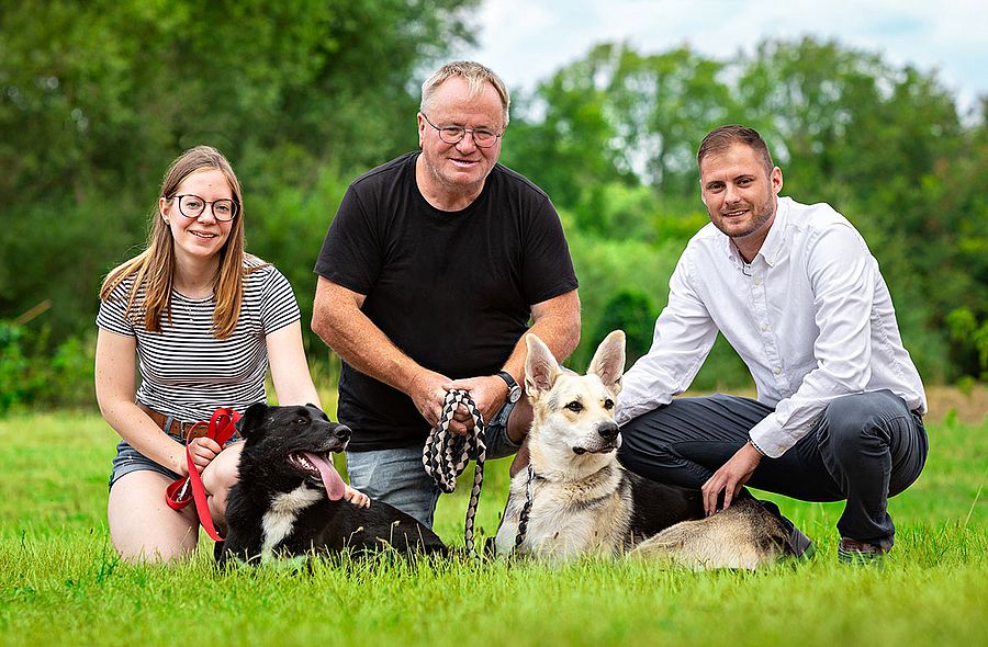 ein foto von 3 menschen knieend mit einem hund auf einer wiese vor einem wald