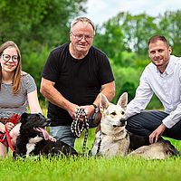 ein foto von 3 menschen knieend mit einem hund auf einer wiese vor einem wald