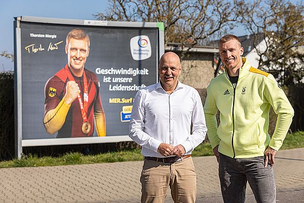 zwei männer posieren vor einer werbetafel