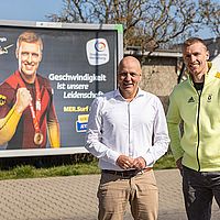 zwei männer posieren vor einer werbetafel