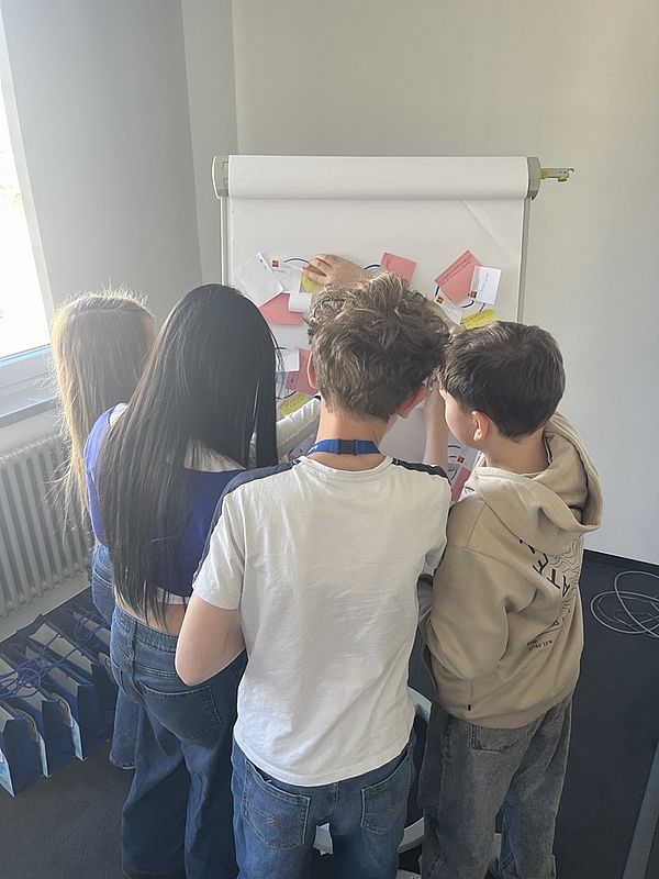 Vier Kinder stehen vor einem Arbeitspapier und bringen Notizen an.