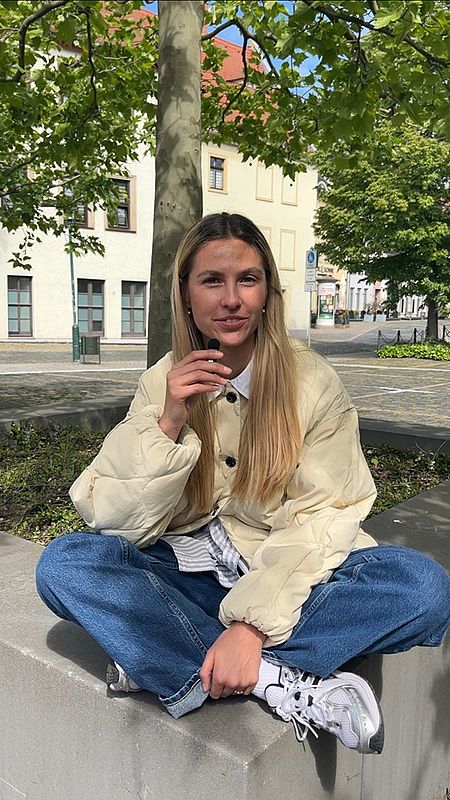 Eine junge blonde Frau mit Mikro in der Hand sitzt auf einer Steinbank unter einem Baum und blickt in die Kamera.