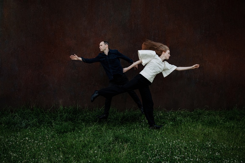 Das Bild zeigt Miles Perkin und Johanna Borchert vor schwarzem Hintergrund auf eine Wiese - sie laufen auseinander.