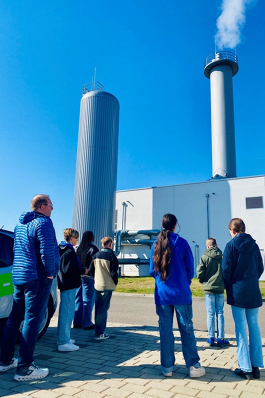 Mädchen und Jungen stehen vor einem Blockheizkraftwerk, das dampft.