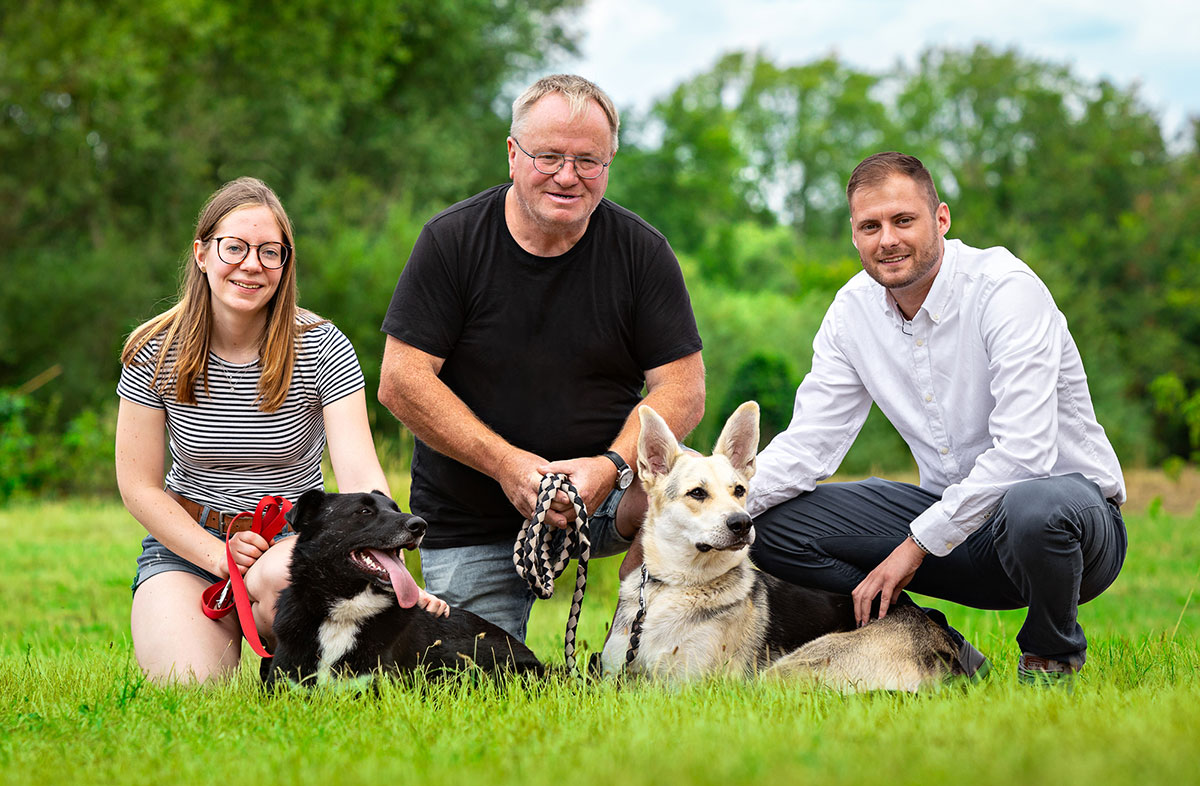 ein foto von 3 menschen knieend mit einem hund auf einer wiese vor einem wald