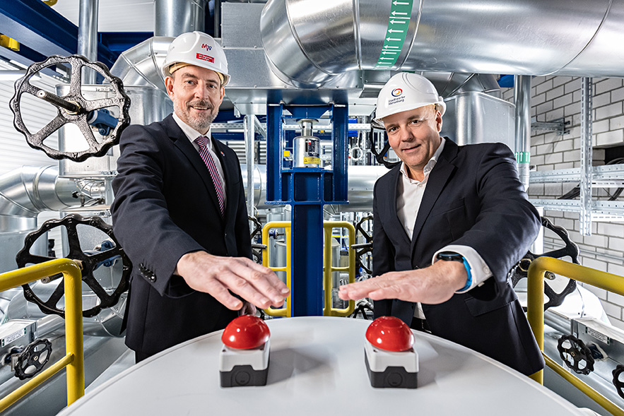 zwei personen im anzug stehen in einer produktionshalle und drücken inen roten busser